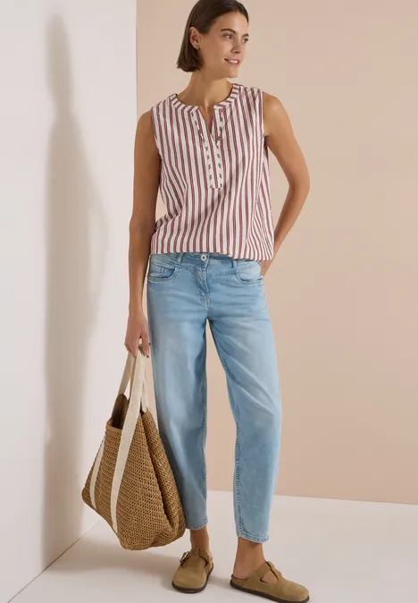 Seersucker Blusentop mit Streifen russet red Frau in gestreiftem ärmellosem Top, hellblauen Jeans und Basttasche vor hellem Hintergrund.