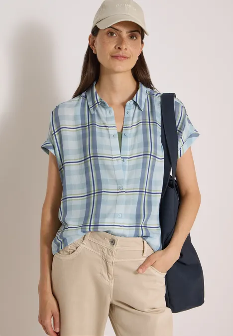Frau trägt kariertes, kurzärmeliges Hemd, beige Hose und Cap, mit Netz Schultertasche, lässig stehend.