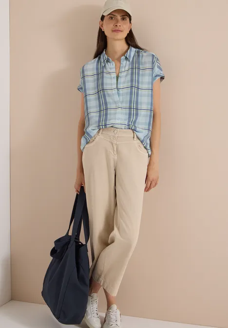 Frau mit kariertem Hemd, beigen Leinenhosen, weißen Sneakers und Cap hält dunkelblaue Tasche.