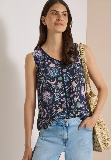 Frau in ärmellosem, dunkelblauem, floralem Top mit hellblauen Jeans und Strohtasche vor hellem Hintergrund.