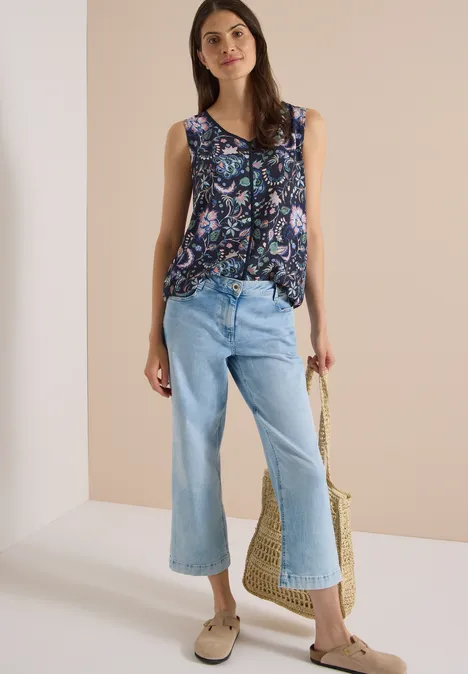 Frau trägt eine bunte, ärmellose Bluse, weite hellblaue Jeans-Culottes und hält eine Strohtasche.