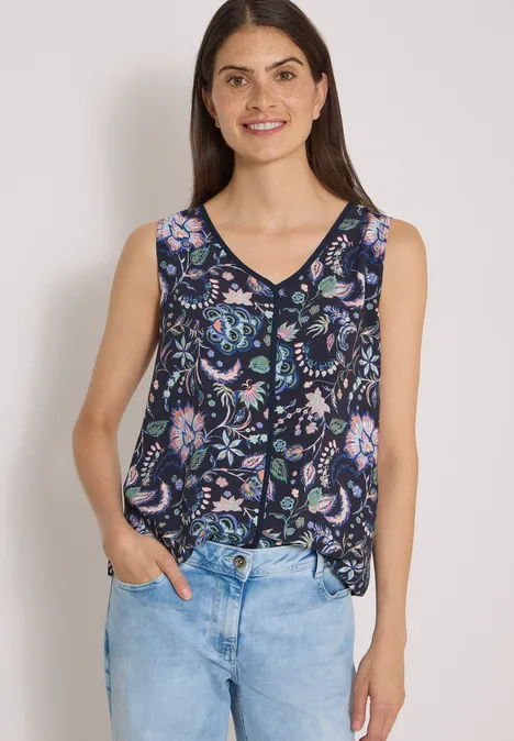 Frau mit dunklen Haaren trägt ärmelloses, geblümtes Top und blaue Jeans, lächelt leicht.
