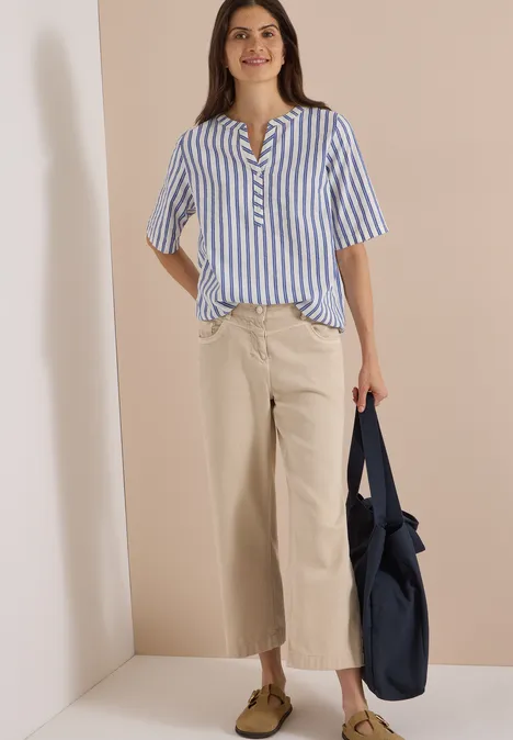 Halbarm Bluse mit Split Neck und Streifen deep river blue Frau vor neutralem Hintergrund, trägt blau-weiß gestreiftes Hemd, beige Culotte-Hose, braune Schuhe, hält navy Tasche.