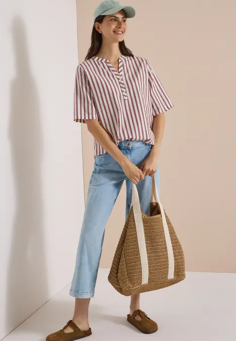 Halbarm Bluse mit Split Neck und Streifen russet red Frau in gestreiftem Top, Jeans und Cap hält eine geflochtene Tasche, steht fröhlich vor neutralem Hintergrund.