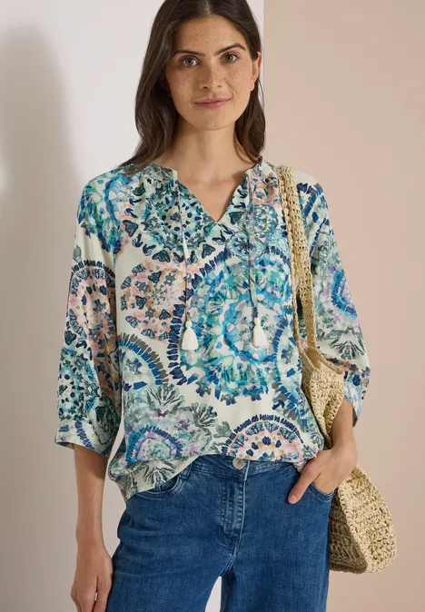 Frau in gemusterter Bluse mit Blau- und Grüntönen, Jeans und Strohtasche