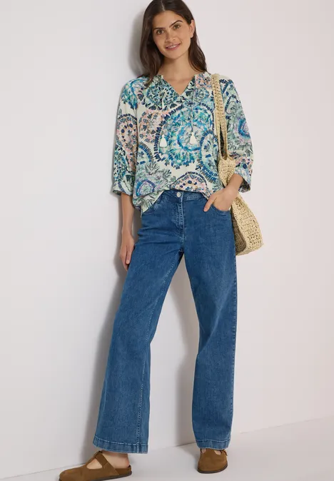 Frau in gemusterter Bluse, breiten Jeans und Stroh-Tasche vor hellem Hintergrund.