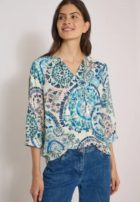 Eine lächelnde Frau trägt eine bunte gemusterte Bluse und blaue Jeans vor neutralem Hintergrund.