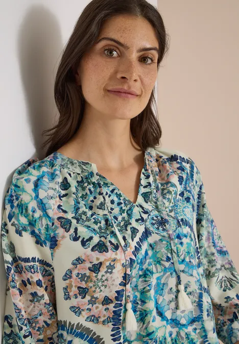 Eine Frau mit dunklen Haaren trägt eine weiße Bluse mit buntem, blau-grünem Blumenmuster.