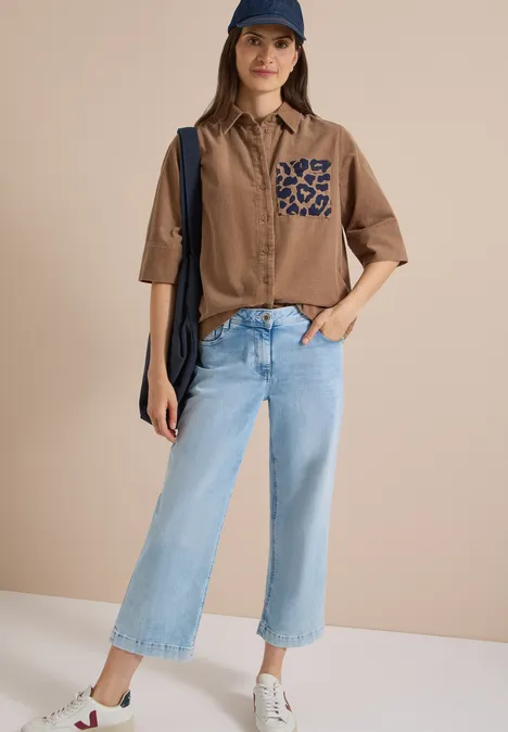 Chemisier à manches courtes en velours côtelé avec poche poitrine creamy mocha Frau mit Cap, braunem Hemd mit Leoparden-Tasche, hellblauen Cropped-Jeans, weißen Sneakern und Schultertasche vor neutralem Hintergrund.