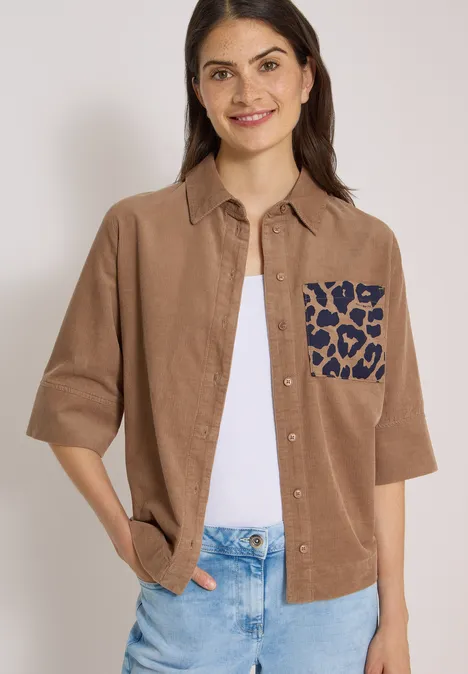 Chemisier à manches courtes en velours côtelé avec poche poitrine creamy mocha Lächelnde Frau in braunem Cordhemd mit blau gefleckter Leoparden-Tasche, weißem Top und Jeans.