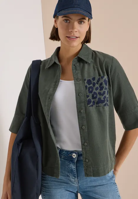 Frau trägt grüne Bluse mit Leopardenmuster-Tasche, weißem Top, Jeans, dunkelblauer Cap und Tasche.