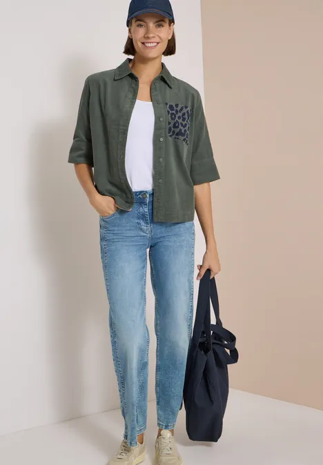 Frau in Jeans, weißem T-Shirt, grüner Bluse, blauer Mütze trägt eine große blaue Tasche.