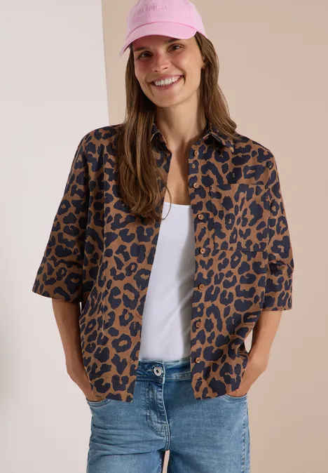 Junge Frau mit rosa Baseballcap, weißem Top, braun-schwarz gemustertem Overshirt und Jeans lächelt in die Kamera.