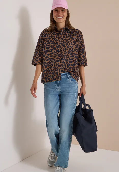 Eine lächelnde Frau mit rosa Cap, Leo-Shirt, Jeans, Sneakers trägt eine blaue Tasche vor hellem Hintergrund.