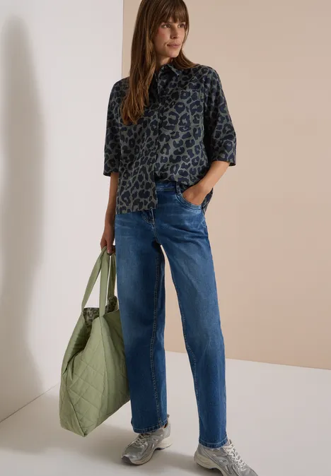 Frau trägt Leopardenmuster-Bluse, blaue Jeans, Sneaker, hält große grüne Tasche