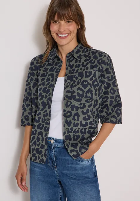Eine lächelnde Frau trägt eine kurzärmelige, grüne Bluse mit Leo-Muster, weißen Top und Jeans.