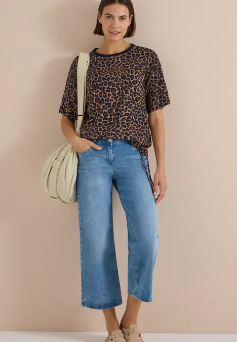 Eine Frau trägt eine Leo-Bluse, weite Jeans-Culotte und helle Tasche vor beigem Hintergrund.
