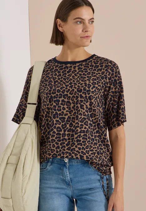 Frau in kurzärmeligem, braun-schwarzem Leo-Shirt mit Jeans und beiger Stepp-Umhängetasche.