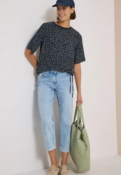 Junge Frau in schwarzem, gemustertem T-Shirt, hellen Jeans, Cap und grüner Stepp-Tasche.