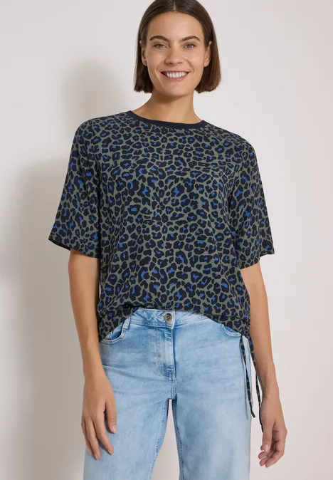 Eine Frau lächelt trägt ein blau-schwarz gemustertes T-Shirt mit Knoten am Saum und helle Jeans vor weißem Hintergrund