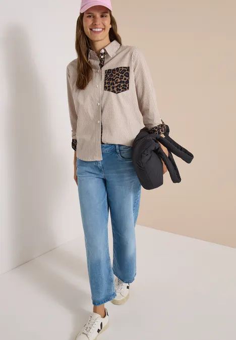 Frau in Jeans, heller Jacke mit Leoparden-Tasche, Cap und Sneakern, trägt eine Tasche mit Stativ.