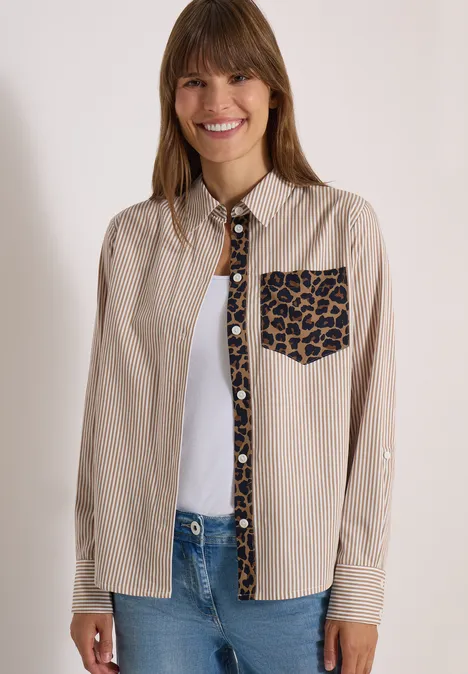 Frau trägt beige Hemd mit Leopardenmuster an Brusttasche und Knopfleiste, darunter weißes Top und Jeans.