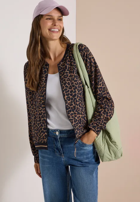 Vrouw met pet, luipaard bomberjack, wit topje, spijkerbroek en groene tas voor een tweekleurige achtergrond.