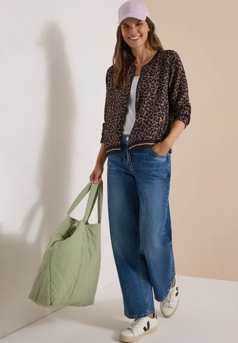 Vrouw draagt luipaardjas, wit topje, blauwe jeans, witte sneakers en roze pet, met grote groene tas.