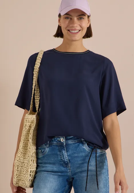 Kurzarm Blusenshirt mit Rundhals urban dark blue Lächelnde Frau in dunkelblauem T-Shirt, Jeans und Baseballcap trägt eine geflochtene Umhängetasche.