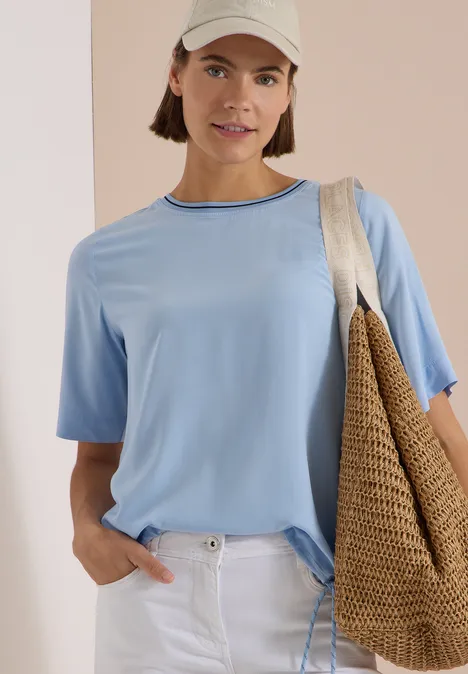 Kurzarm Blusenshirt mit Rundhals easy blue Frau in hellblauem T-Shirt, weißer Hose und Cap, mit Strohtasche vor hellem Hintergrund.