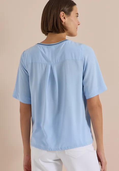 Kurzarm Blusenshirt mit Rundhals easy blue Frau von hinten in hellblauem T-Shirt mit kurzen Ärmeln und weißer Hose vor beigem Hintergrund.