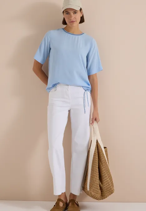 Kurzarm Blusenshirt mit Rundhals easy blue Frau in hellblauem T-Shirt, weißer Hose, Strohtasche und Cap vor beigem Hintergrund.