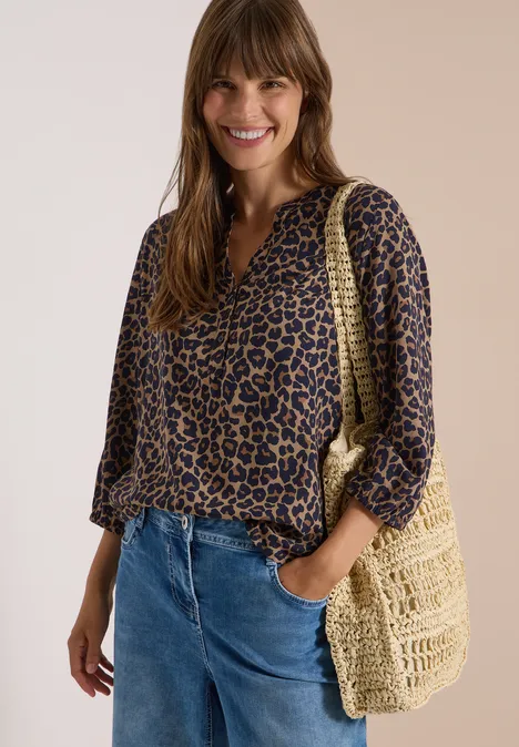 Frau mit Leoparden-Print-Bluse, blauen Jeans und geflochtener Tasche lächelt.