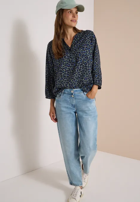 Tunique léopard à manches 3/4 et col fendu dried olive green Frau trägt blau gemusterte Bluse, helle Jeans, weiße Sneaker und grüne Kappe.