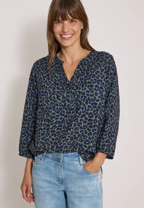 Tunique léopard à manches 3/4 et col fendu dried olive green Frau trägt Jeans und dunkelblaue Bluse mit Leopardenmuster und V-Ausschnitt, lässig.