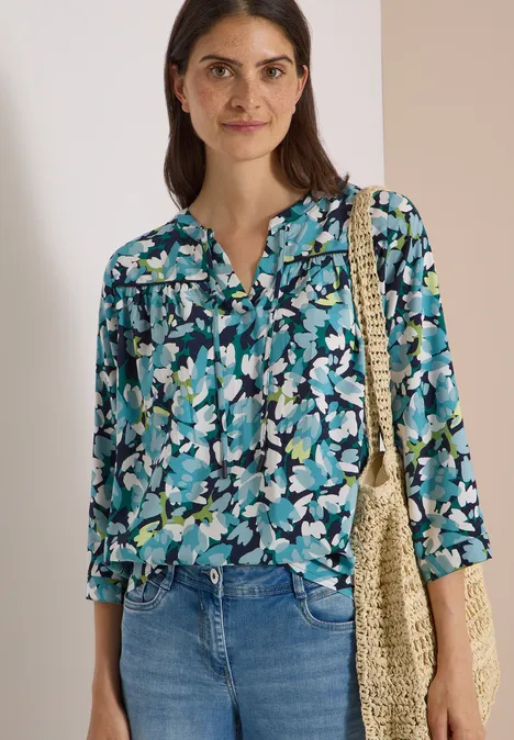 3/4-Arm Tunika mit Split Neck und Print urban dark blue Frau trägt geblümte Bluse in Blaugrün, Jeans und einen gehäkelten Stoffbeutel.