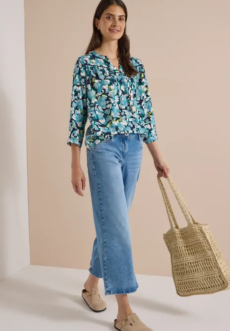 3/4-Arm Tunika mit Split Neck und Print urban dark blue Frau in blau-grün geblümter Bluse, weiter Jeans, beigen Schuhen und Korbtasche vor hellem Hintergrund.