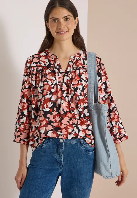 3/4-Arm Tunika mit Split Neck und Print russet red Frau in floraler Bluse und Jeans, lächelt und trägt eine Schultertasche vor hellem Hintergrund.