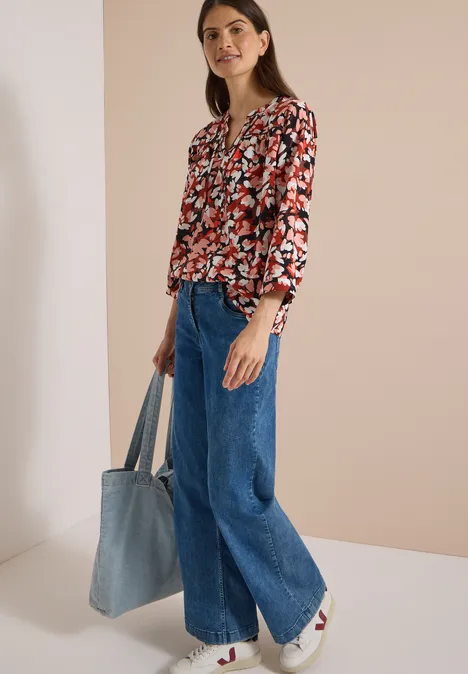 3/4-Arm Tunika mit Split Neck und Print russet red Frau in floraler Bluse und weitem Jeans, weiße Sneaker, hält große Denim-Tasche vor hellem Hintergrund.