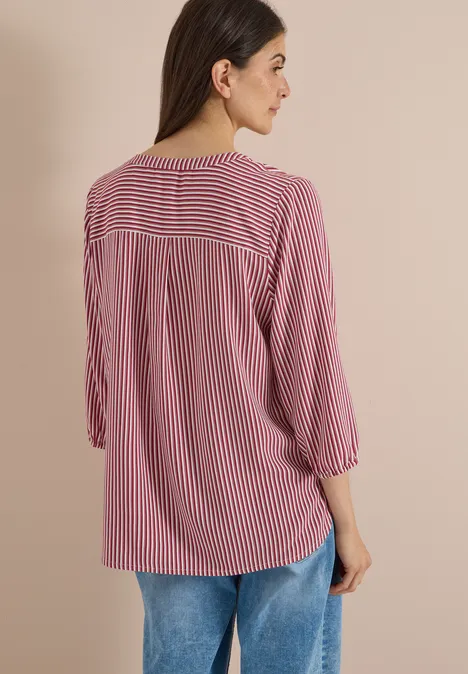 3/4-Arm Bluse mit Split Neck und Streifen russet red Frau von hinten trägt rosarote, weiß gestreifte Bluse und blaue Jeans vor beigem Hintergrund.