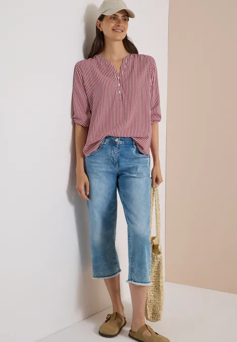 3/4-Arm Bluse mit Split Neck und Streifen russet red Frau lehnt lässig an Wand, trägt lila Bluse, weite Jeans, Cap, Sandalen und Basttasche.