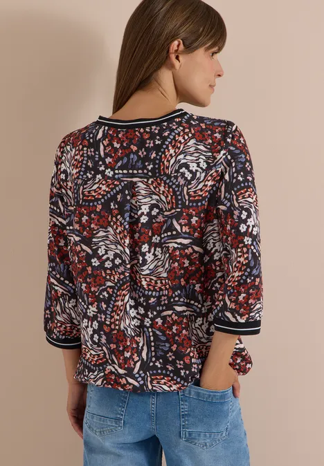 Frau von hinten in rot-blau gemusterter, dreiviertelärmeliger Bluse und hellen Jeans.