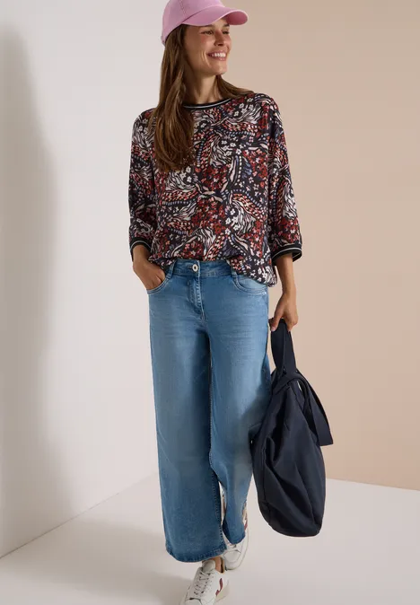 Frau in gemustertem Langarmshirt, weiter Jeans, rosa Cap, weissen Sneakern, hält navy Tasche und lächelt.