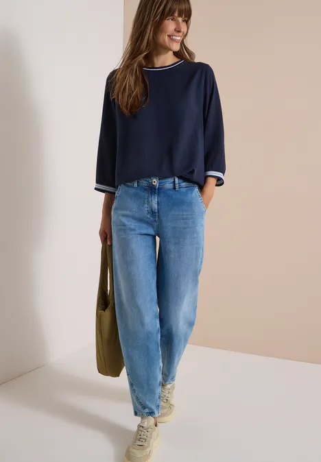 Frau in blauem Pullover und Jeans trägt Tasche und Sneaker vor beigem Hintergrund.