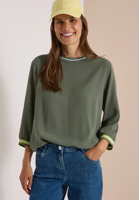 Frau trägt olivgrünes Sweatshirt und Jeans, gelbe Kappe, lächelt locker