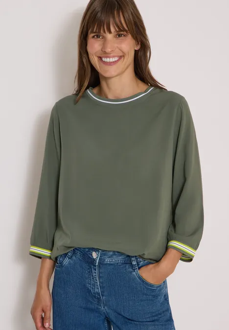 Frau lächelt in legerem Outfit: olivgrünes Sweatshirt mit Streifendetails, Dreiviertelärmel und Jeans.
