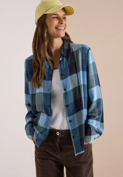 Femme souriante en chemise à carreaux bleue, T-shirt blanc, casquette jaune et pantalon marron.