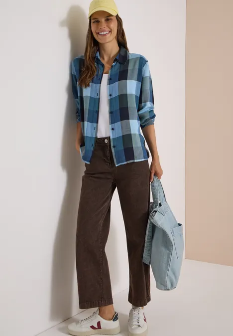 Femme en chemise à carreaux bleu, t-shirt blanc, pantalon marron, baskets et casquette jaune, sac en jean.