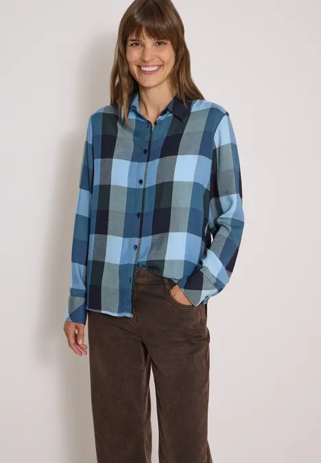 Une femme souriante porte une chemise à carreaux bleue et un pantalon marron sur fond clair.