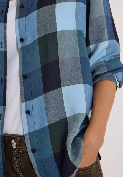 Buste vêtu d’une chemise à carreaux bleue et d’un t-shirt blanc, main dans la poche d’un pantalon marron.